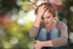 Depressed mum sitting in a park curled with one hand hugging her legs and the other hand touching her head