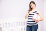 Pregnant Woman on striped shirt holding a piggy bank beside a white crib
