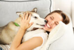 Dog licking a happy woman in whit shirt on the lips on the couch.